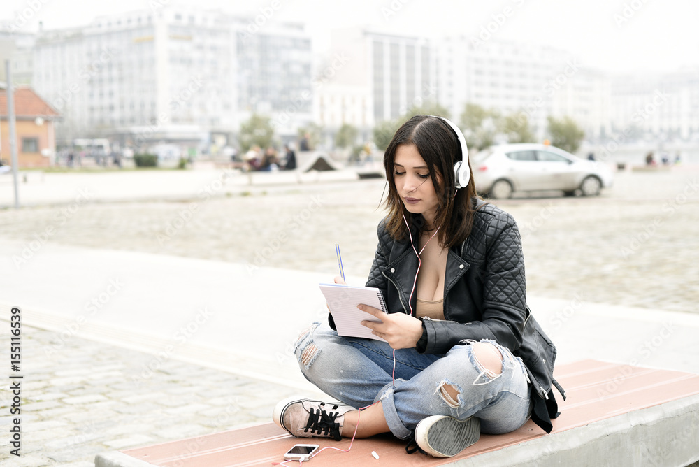 Fototapeta premium Young woman listening music from a smart phone and writing notes