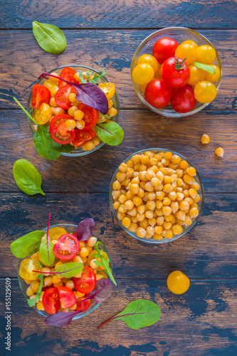 Fototapeta Naklejka Na Ścianę i Meble -  Healthy vegetarian salad with chickpeas, red and yellow cherry tomatoes and green leaves. Small glass bowls on the rustic dark wooden table, top view.