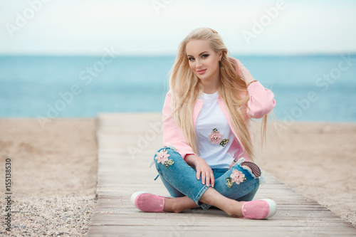 Beautiful autumn woman wear pink jacket posing near the seaa outdoors. Beauty happy long hairl blonde girl smiling at beach. Autumn  style fashion model photo