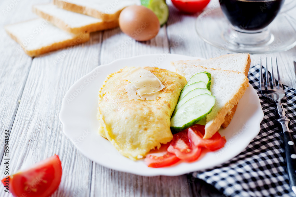 Hearty and tasty breakfast, traditional in the hotel, omelette from chicken eggs with cheese, fresh vegetables - cucumber and tomato, white toast and black coffee on a light wooden background