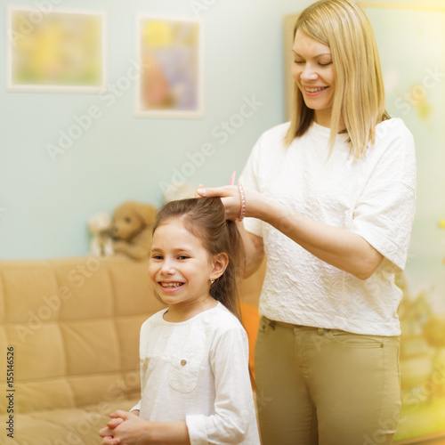 Mother is braiding her young daughter's hair standing in the children room.