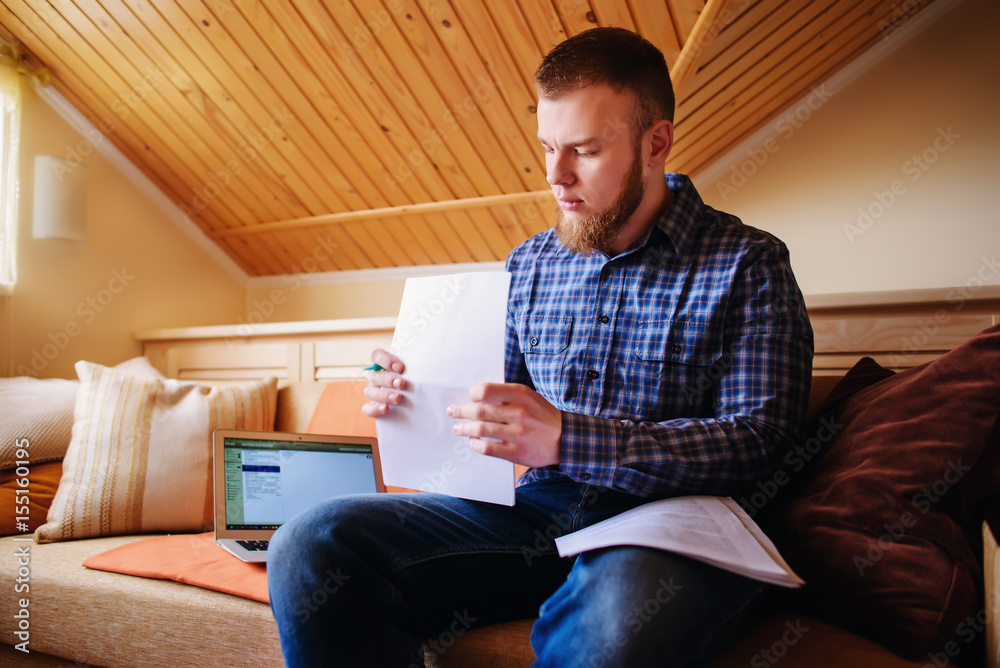 Young man studying from home sitting on a sofa holding documents while working on a laptop computer Young man studying from home sitting on a sofa holding documents while working on a laptop computer