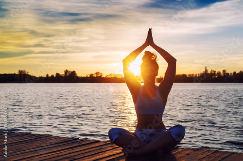 Young woman doing yoga exercises on the lake beach at sunset