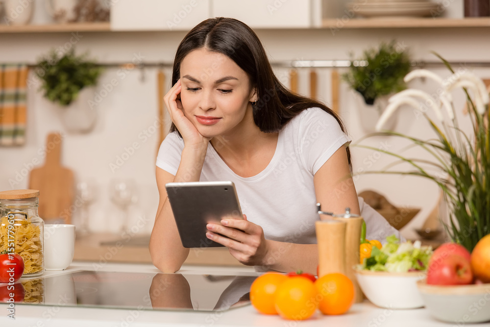 Young Woman standing in kitchen with tablet computer and looking recipes.
