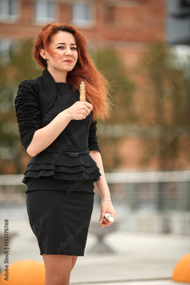 Vape teenager. Young pretty white girl with red curly hair and modern haircut in black dress vaping an electronic cigarette opposite city background. Vape teenager. Young pretty white girl with red curly hair and modern haircut in black dress vaping an electronic cigarette opposite city background.