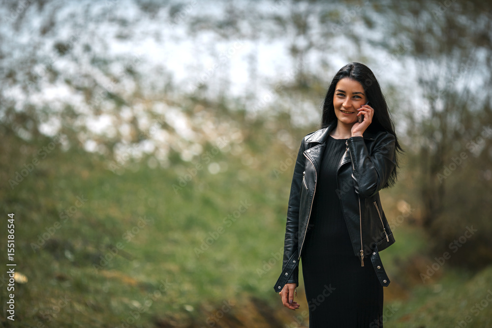 Young pretty white girl with black hair and in black clothes talking on the phone against the edge of the forest with snow in the spring. Young pretty white girl with black hair and in black clothes talking on the phone against the edge of the forest with snow in the spring.