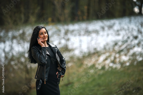 Young pretty white girl with black hair and in black clothes talking on the phone against the edge of the forest with snow in the spring.