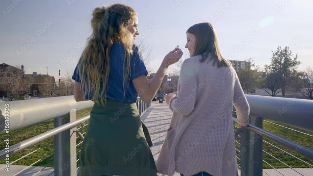 Two Teens Walk Away, Across A Bridge In A City Park, Girl Shares Her Headphones With Her Friend And They Sing And Dance Two Teens Walk Away, Across A Bridge In A City Park, Girl Shares Her Headphones With Her Friend And They Sing And Dance