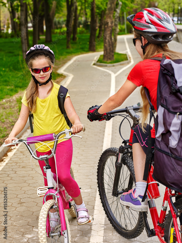 Fototapeta premium Bicycle path and sign with children. Girls wearing helmet with rucksack ciclyng ride. Kids are on white bike lane. Alternative to urban transport. Oncoming traffic. People support healthy lifestyle.