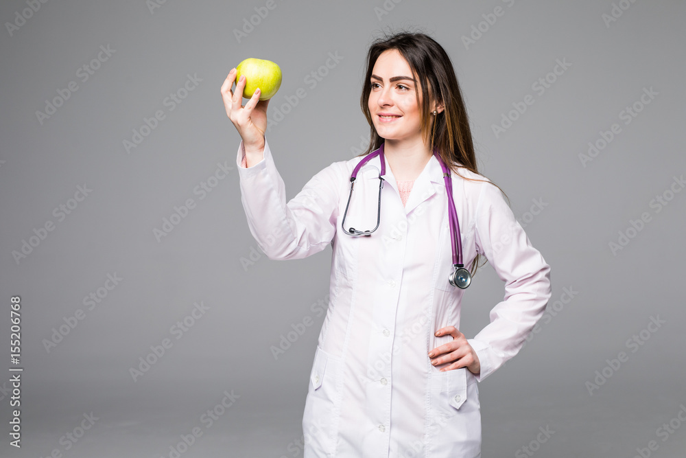 Cheerful cute young woman doctor standing and holding an apple on grey Cheerful cute young woman doctor standing and holding an apple on grey