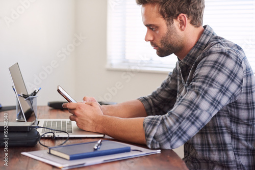 Young adult caucasian man busy using his cellphone while seated at his beautiful wooden desk at home, wearing a checked shirt.