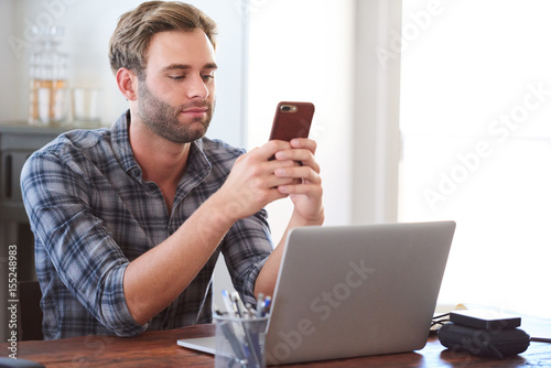 Young man smiling slightly as he flirts via text message on his phone while seated at his desk behind his laptop, procrastinating for as long as possible before getting back to work. photo