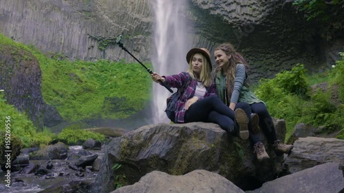 Friends Sit On A Rock And Use A Gopro To Take Selfies Together, With Waterfall In Background
