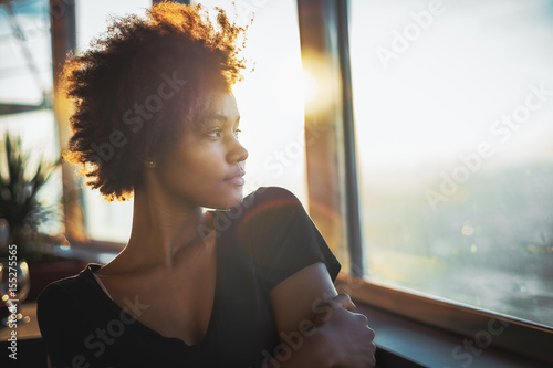 Curly charming young afro american tourist female in black dress is sitting in luxury restaurant of cruise ship near window and thoughtfully looking out the big window on ocean and warm sunset horizon photo