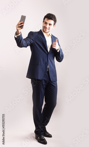 Handsome bearded young business man taking selfie smiling. Full body length portrait isolated over grey studio background.