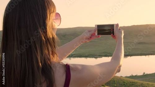 Close up from the back of girl or woman taking photo of sunset lake view landscape with her iphone
