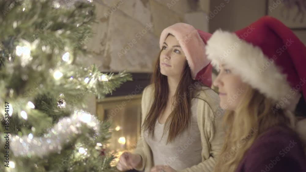 Teen Girls, Wearing Santa Hats, Decorate A Christmas Tree By A Cozy Fire Teen Girls, Wearing Santa Hats, Decorate A Christmas Tree By A Cozy Fire