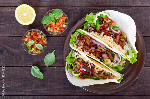 Tacos is a traditional Mexican dish. Tortilla stuffed with chicken, bell and hot peppers, beans, lettuce, cheese, blue onion with salsa sauce on dark wooden background. Top view.
