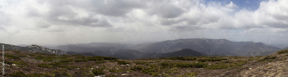 Panoramic landscape with peaks of mountains, hills, forest and sky with contrasting clouds Panoramic landscape with peaks of mountains, hills, forest and sky with contrasting clouds