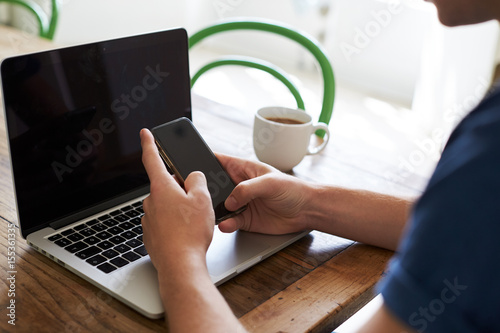 Man Sending Text Message On Mobile Phone Whilst Using Laptop