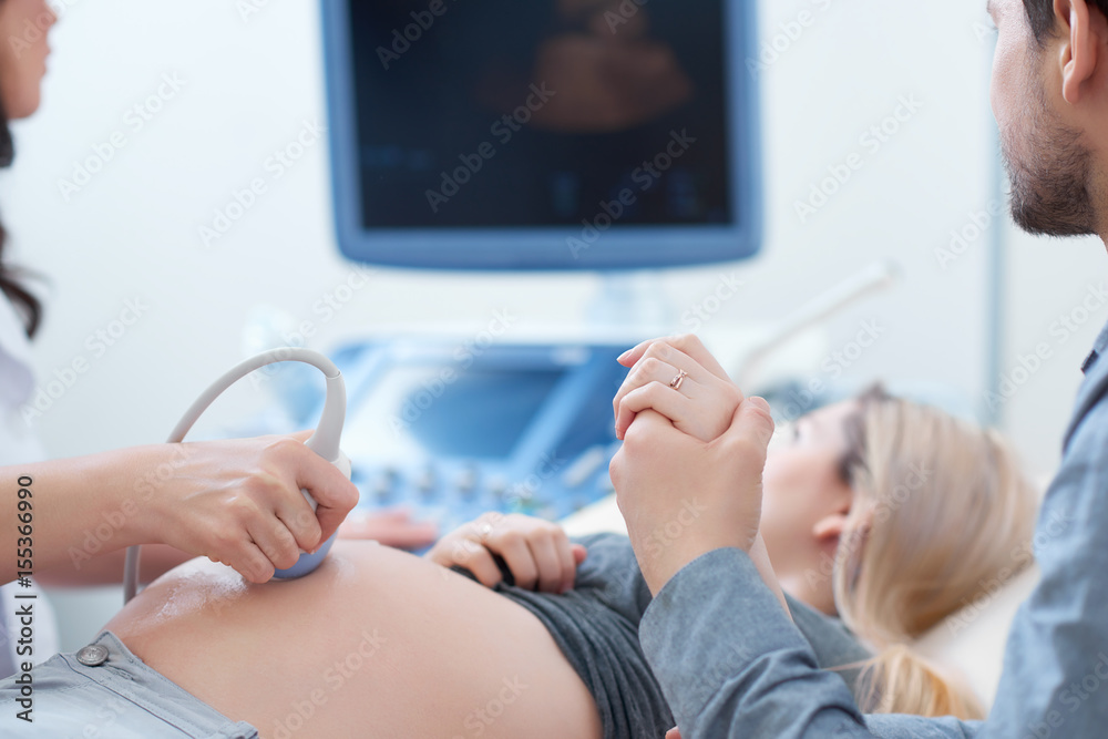 Female doctor touching stomach by tool of woman who lying,showing baby at screen of computer. Professional doctor using ultrasound equipment screening of pregnant woman. Husband holding hand his wife. - obrazy, fototapety, plakaty 