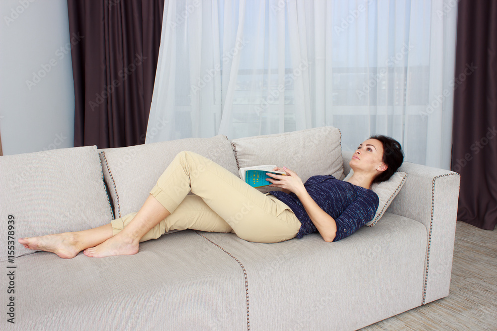 Smiling beautiful brunette relaxing on the couch and reading a book in the living room Smiling beautiful brunette relaxing on the couch and reading a book in the living room