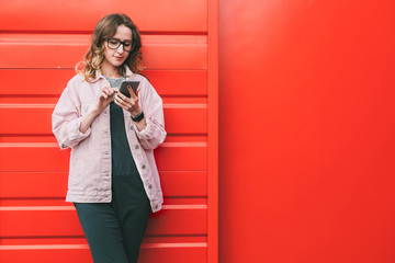 Businesswoman in glasses stands outside on red metal wall background,looking at screen of phone,uses smartphone.