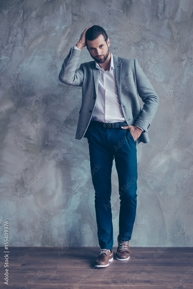 Full length portrait of classy young bearded business man, standing on gray concrete background. He looks spectacular! in a suit, fixing his hairstyle