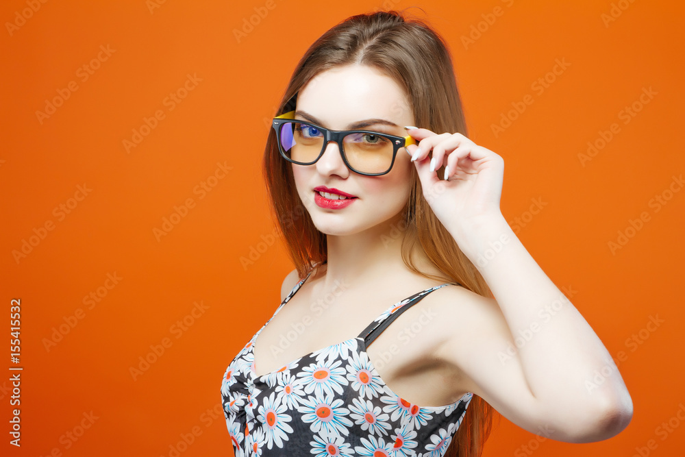 Portrait of Beautiful Female Model in Eyeglasses on Orange Background. Attractive Brunette with Sensual Lips and Dress with Flowers is Posing in Studio. Portrait of Beautiful Female Model in Eyeglasses on Orange Background. Attractive Brunette with Sensual Lips and Dress with Flowers is Posing in Studio.