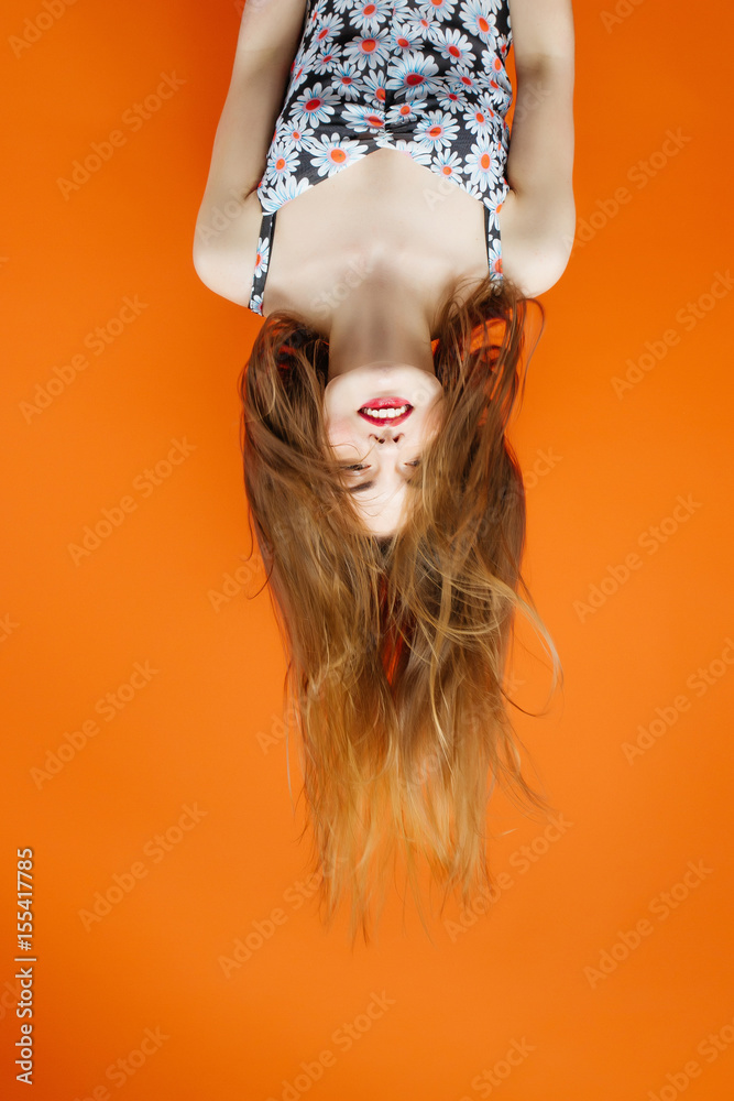 Portrait of Brunette with Long Hair Wearing Dress with Flowers Upside Down in Studio on Orange Background. Hair Style Concept.