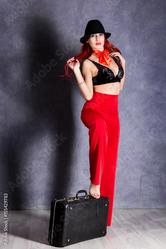 Fototapeta Naklejka Na Ścianę i Meble -  woman in red pants and black hat with big boobs hold retro suitcase