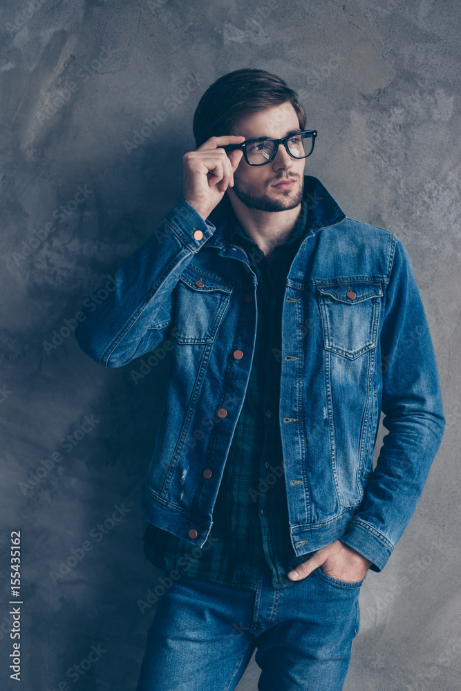 Autumn. Young smart ponder bearded brunette guy with bristle in casual jeans outfit is standing on a concrete wall`s background, wearing glasses and touching them