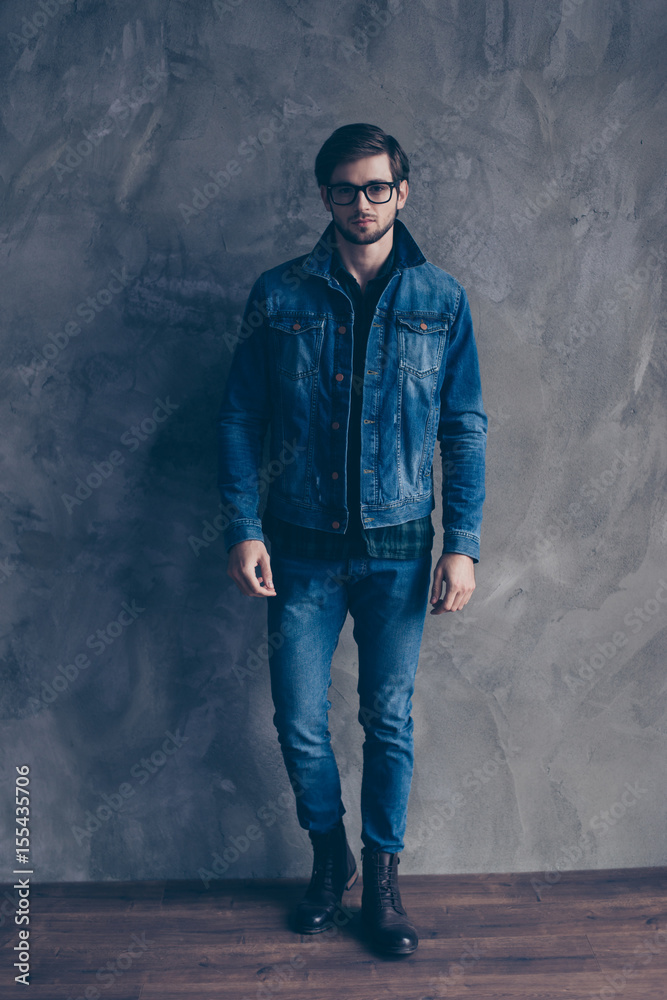 Autumn. Young serious bearded guy in casual jeans outfit is standing on a concrete wall`s background, wearing glasses and look pensive Autumn. Young serious bearded guy in casual jeans outfit is standing on a concrete wall`s background, wearing glasses and look pensive