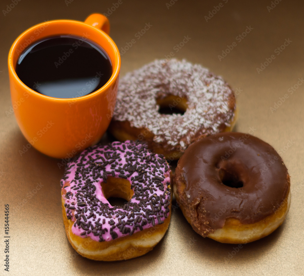 Close up of a delicious donuts with chocolate, coconut and pink glazed with some chips of chocolate, with an orange cup of coffe in a wooden background, top view
