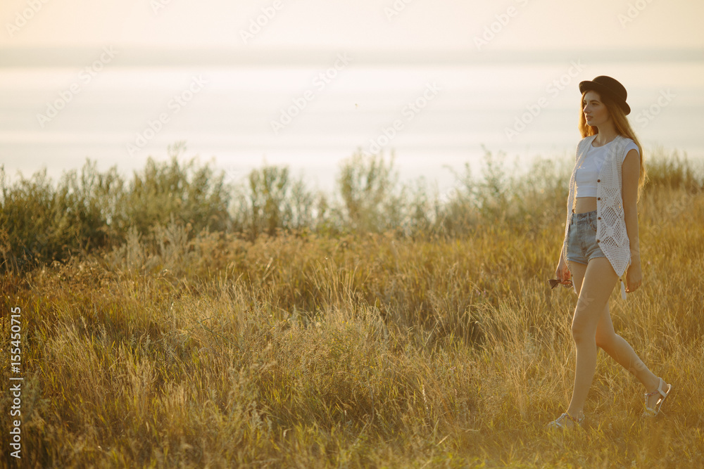 Young pretty woman wearing hat dressed in boho style clothes walking on the nature near river during beautiful sunset