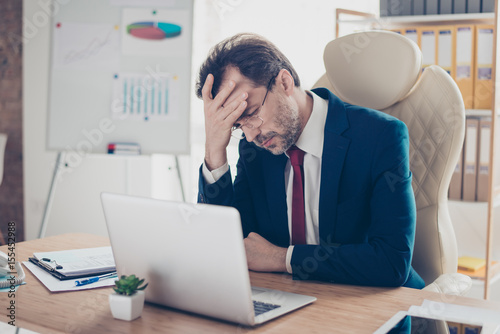 Depressed entrepreneur realtor is holding his head with hand, he is tired and frustrated, sitting in the office at his work place