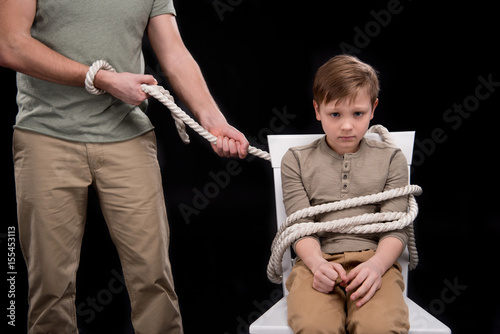 Obraz na plátně Cropped shot of father with rope tied little son sitting on chair, family proble