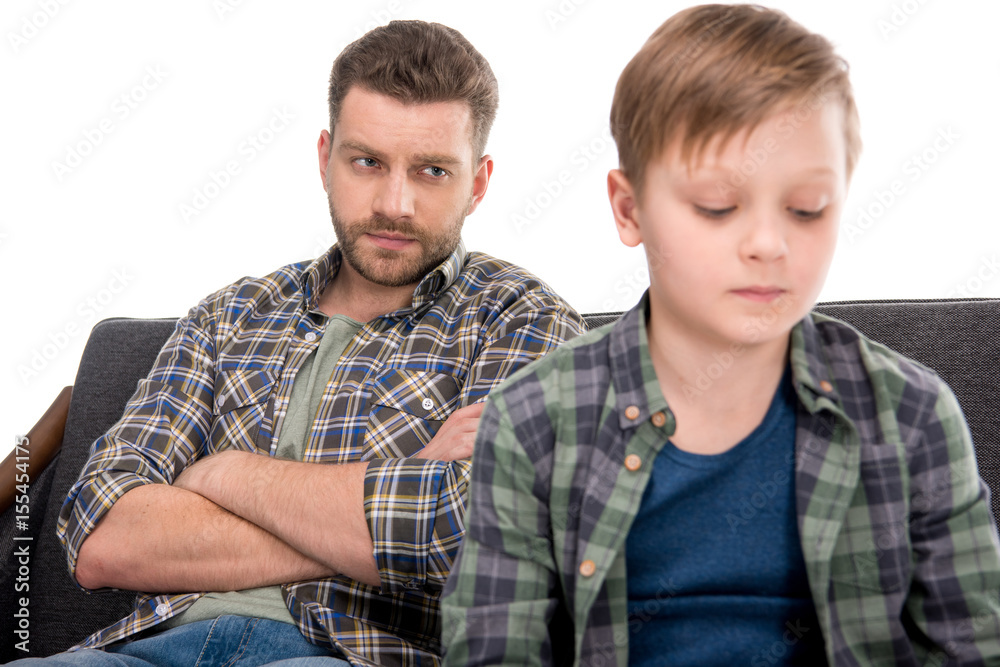 Serious father with crossed arms looking at upset little boy, family problems concept Serious father with crossed arms looking at upset little boy, family problems concept