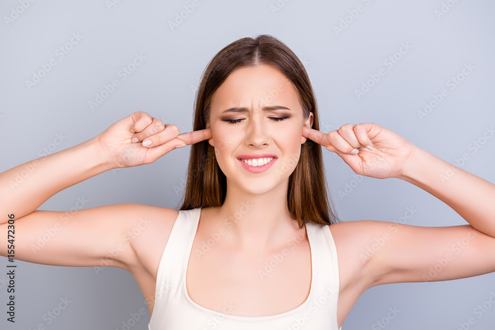 Portrait of tired young cute girl, closing her ears with fingers. She is with closed eyes also, in a white casual singlet on pure background Portrait of tired young cute girl, closing her ears with fingers. She is with closed eyes also, in a white casual singlet on pure background