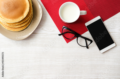 Wooden table with pancakes, cup of milk, glasses and phone