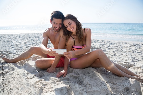 Smiling young couple taking selfie while sitting at beach photo