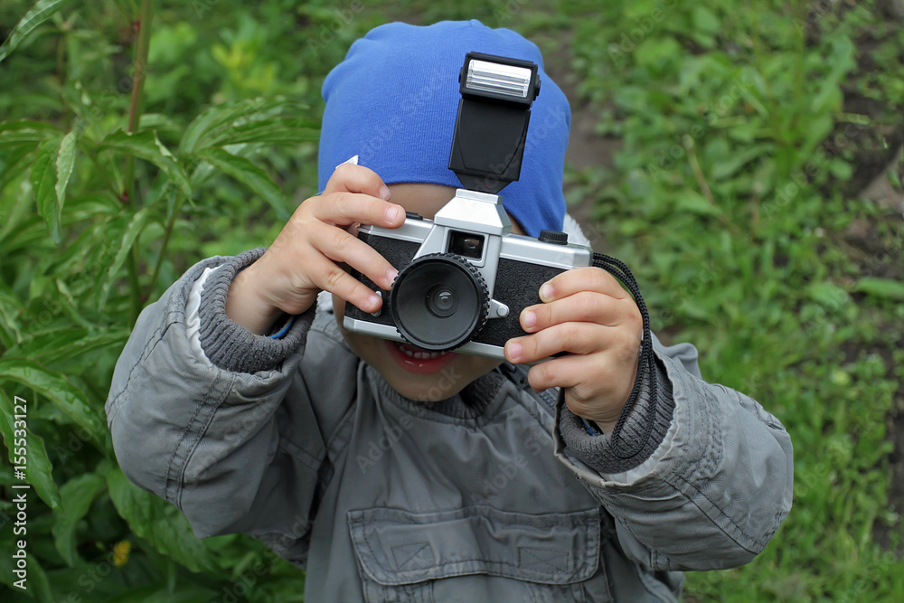 A little boy in a jacket and a blue hat takes pictures of the photographer. Young photographer at work. The kid looks through the lens to the camera A little boy in a jacket and a blue hat takes pictures of the photographer. Young photographer at work. The kid looks through the lens to the camera