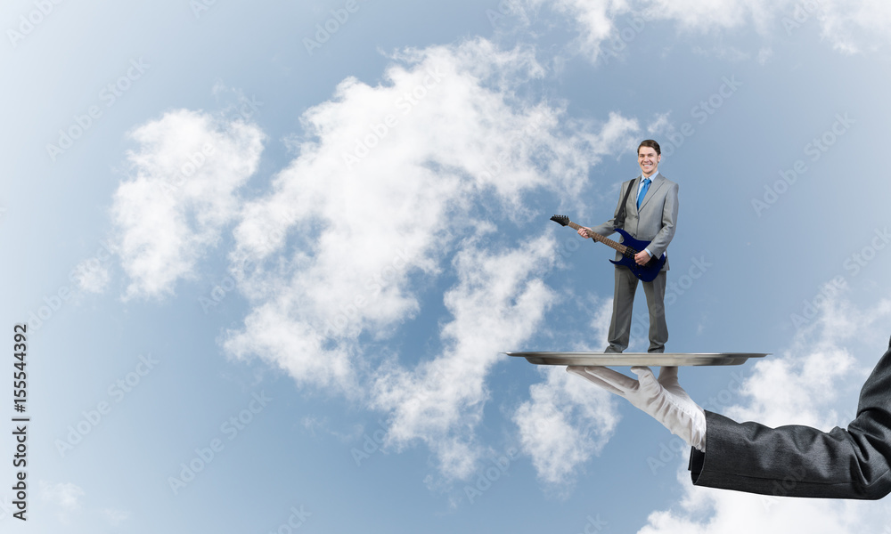 Businessman on metal tray playing electric guitar against blue sky background