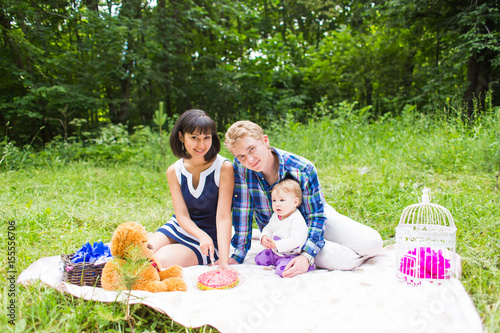 Happy young mother and father with their baby daughter relaxing on a blanket in a park celebrating with birthday cake