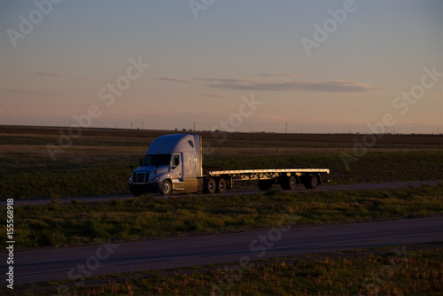A white Freightliner Cascadia pulls an empty flatbed trailer down a rural US Highway during sunset hours. All visible trademarks and markings have been removed. 