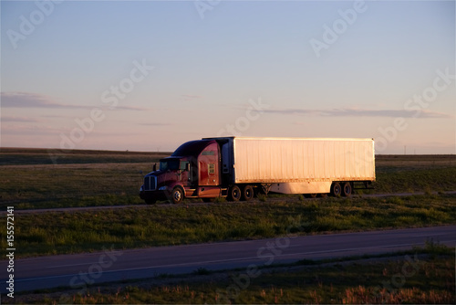 A red or Maroon Kenworth Semi truck pulls a white trailer down a rural US Highway during sunset. All visible markings and trademarks have been removed.  photo