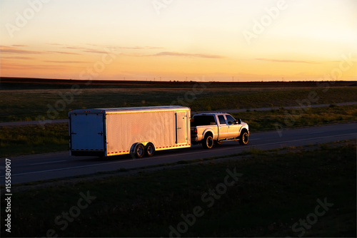 A white pickup truck pulls a small white box trailer down a rural highway during sunset hours. All visible markings and trademarks have been removed. photo