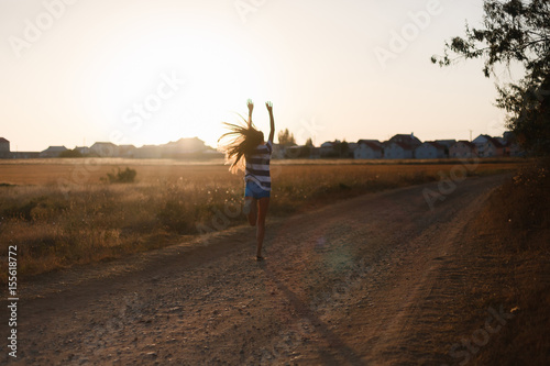 The girl with long hair running and enjoys raising his hands up on the sand road at sunset along the field with dry grass