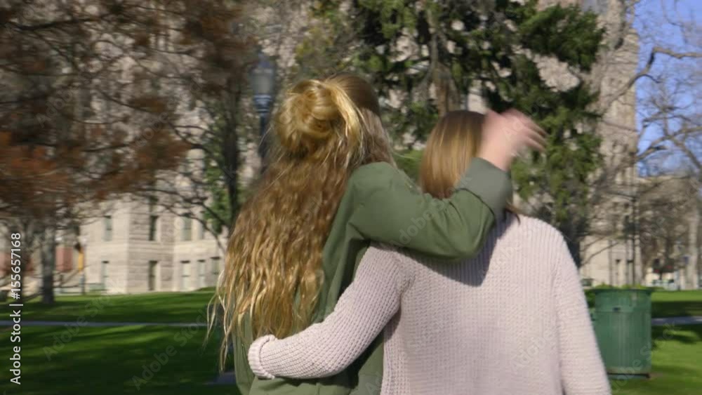 Playful Teens Walk With Arms Around Each Other Through City Park, They Push Each Other And Laugh Playful Teens Walk With Arms Around Each Other Through City Park, They Push Each Other And Laugh