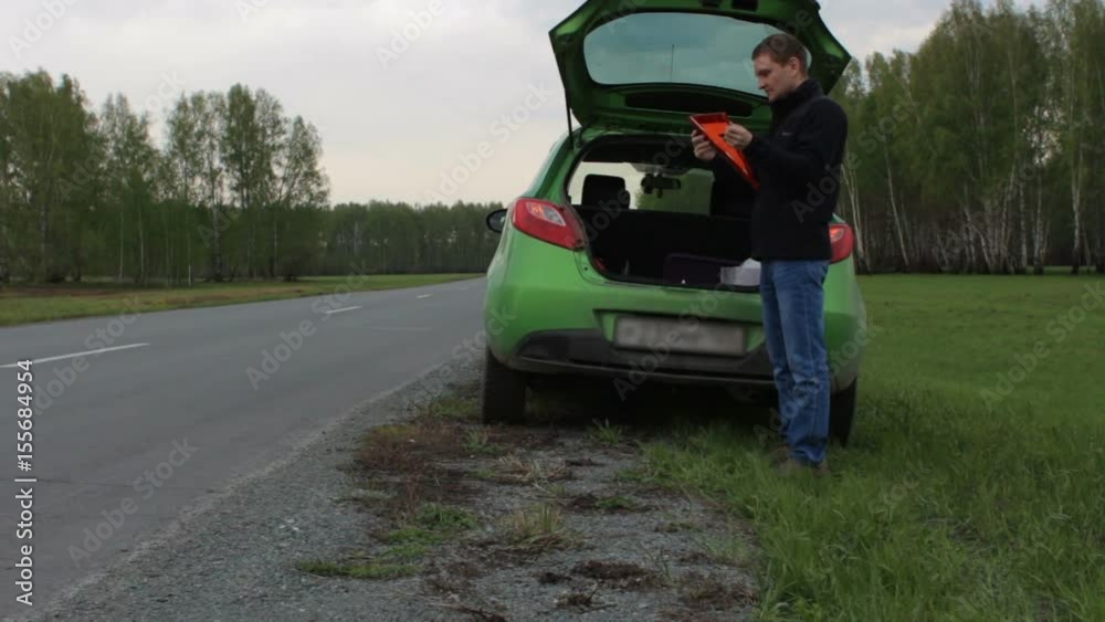 Young man with broken car on the rural road takes triangle from car trunk Young man with broken car on the rural road takes triangle from car trunk